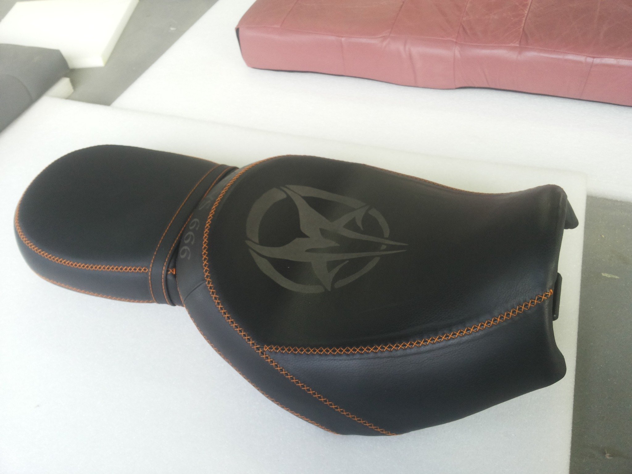 Czarne, skórzane siedzenie motocyklowe z pomarańczowymi przeszyciami i wygrawerowanym wzorem na białym podłożu, obok fragmentu innego siedzenia.