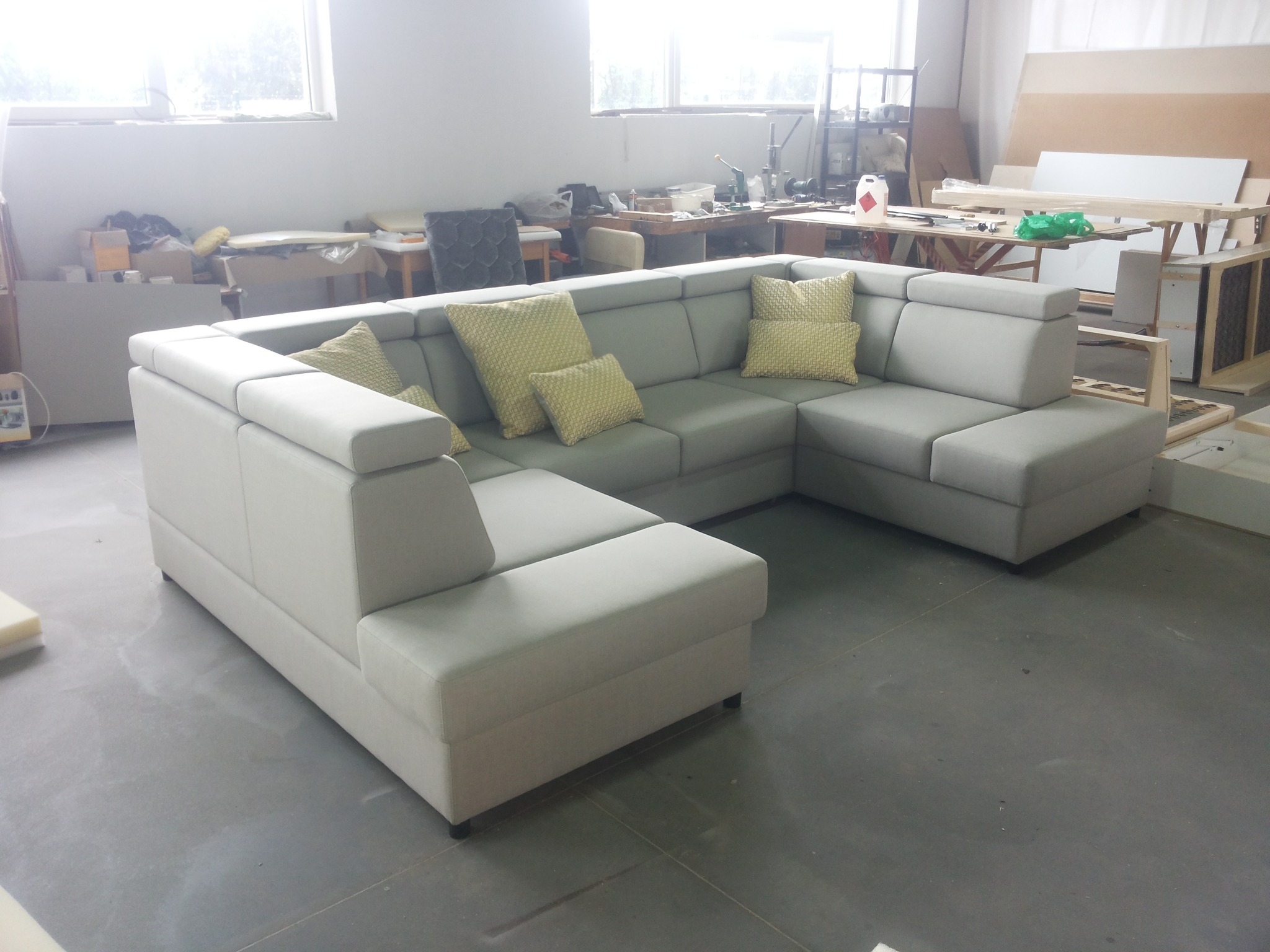 Szara, modułowa sofa narożna z jasnożółtymi poduszkami w warsztacie, widoczne narzędzia i materiały w tle.