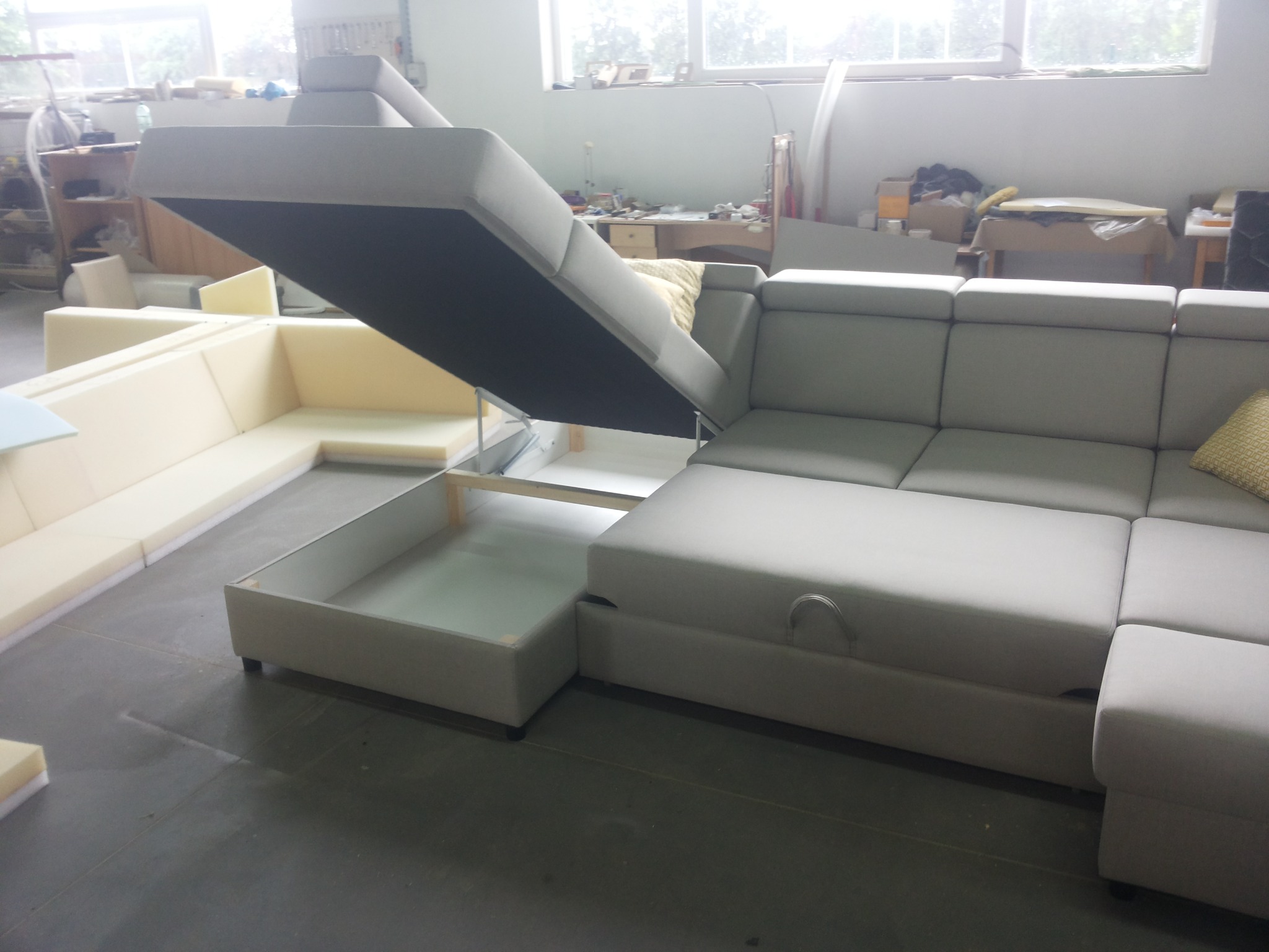 Szary narożnik z podnoszonym siedziskiem ukazującym schowek; widoczne elementy konstrukcyjne i piankowe wypełnienie, w tle warsztat tapicerski.