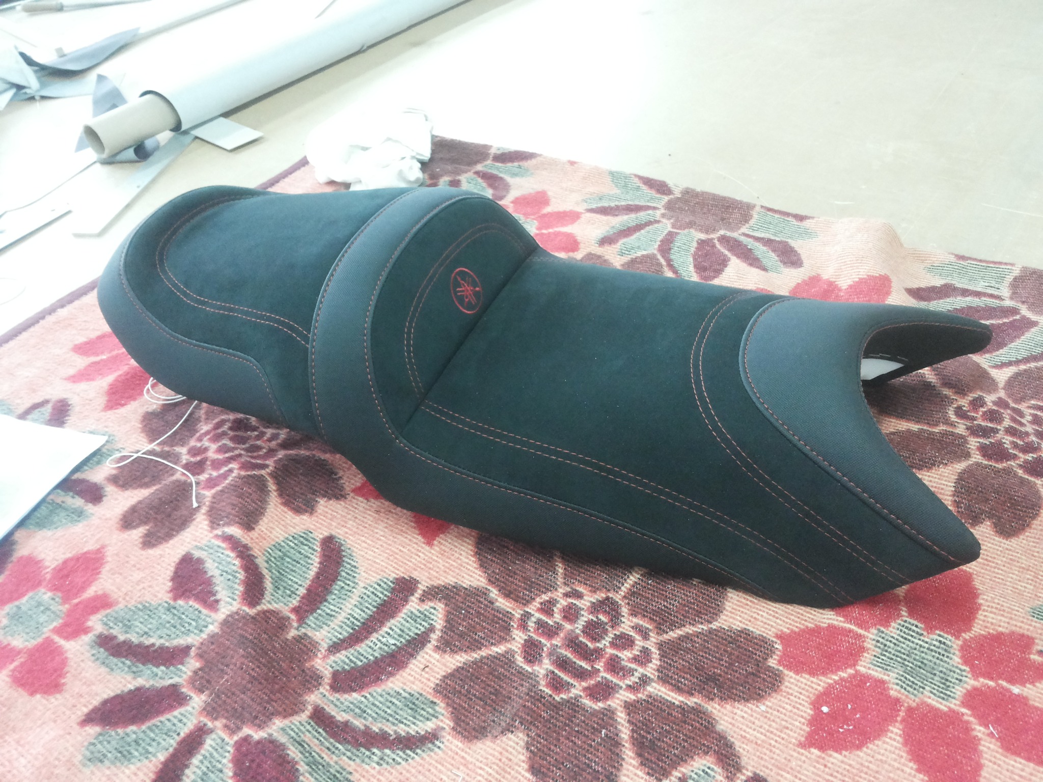 Czarne, tapicerowane siedzisko motocyklowe Yamaha z czerwonym logo, leżące na wzorzystym kocu.