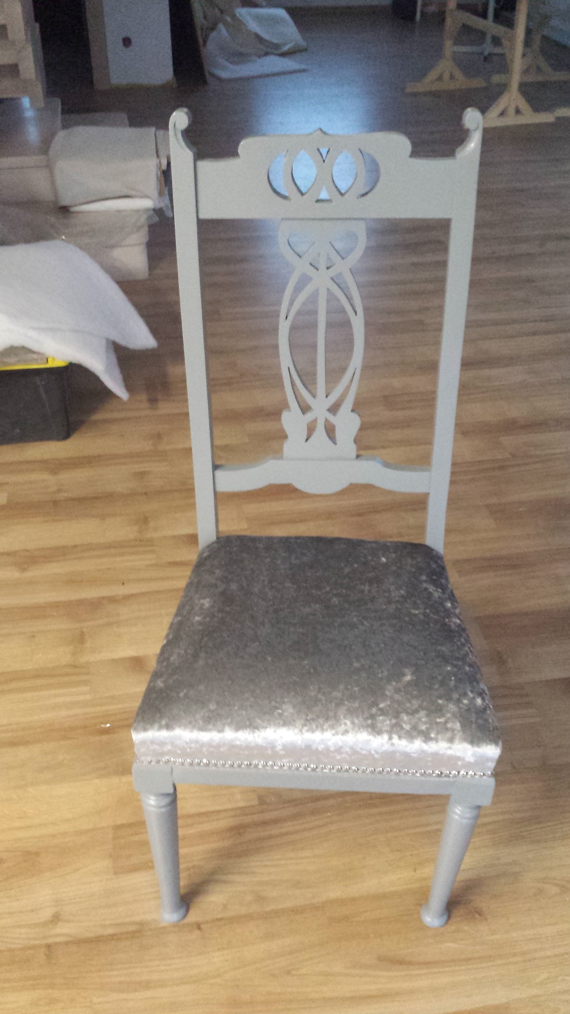 Odnowione krzesło w stylu vintage, pomalowane na szaro, z siedziskiem obitym srebrzystym aksamitem i ozdobnymi pineskami. W tle widoczne elementy wyposażenia warsztatu.