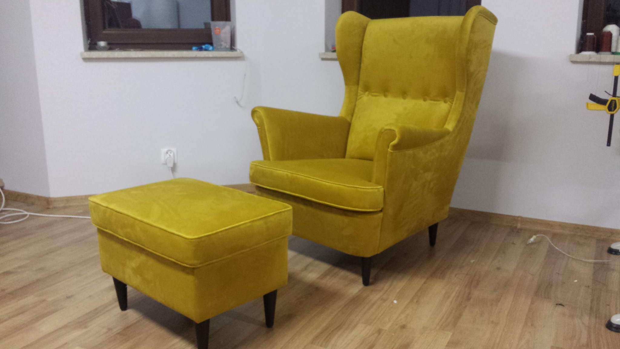 Żółty fotel typu uszak z podnóżkiem na drewnianej podłodze, widoczne okno i narzędzia tapicerskie w tle.