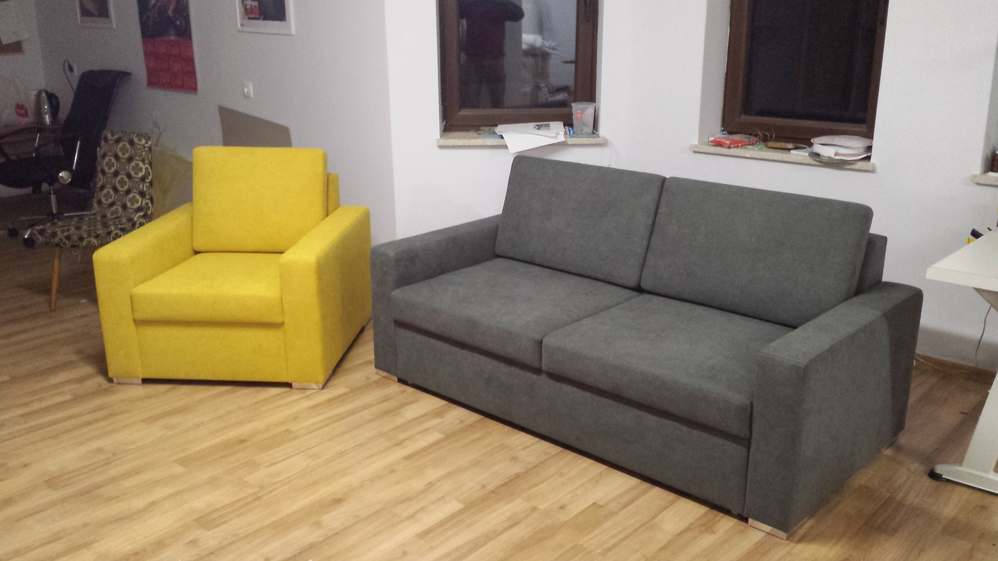 Szary narożnik i żółty fotel w minimalistycznym salonie z drewnianą podłogą i oknami.