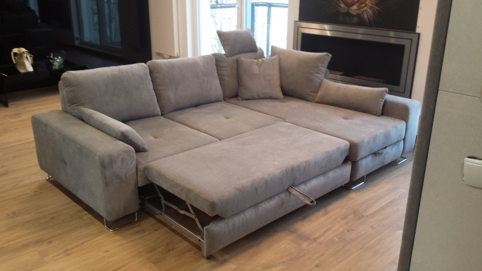 Rozłożona, szara, narożna sofa z wysuwanym siedziskiem i poduszkami dekoracyjnymi na drewnianej podłodze, widok z boku.