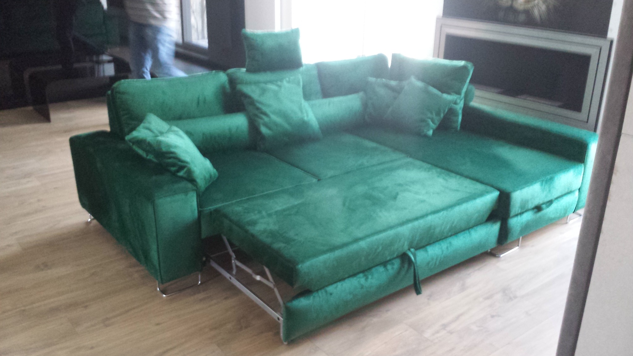 Rozłożona narożna sofa w odcieniach butelkowej zieleni z poduszkami, widoczne metalowe nóżki i mechanizm rozkładania, w tle fragment okna i dekoracji ściennej.