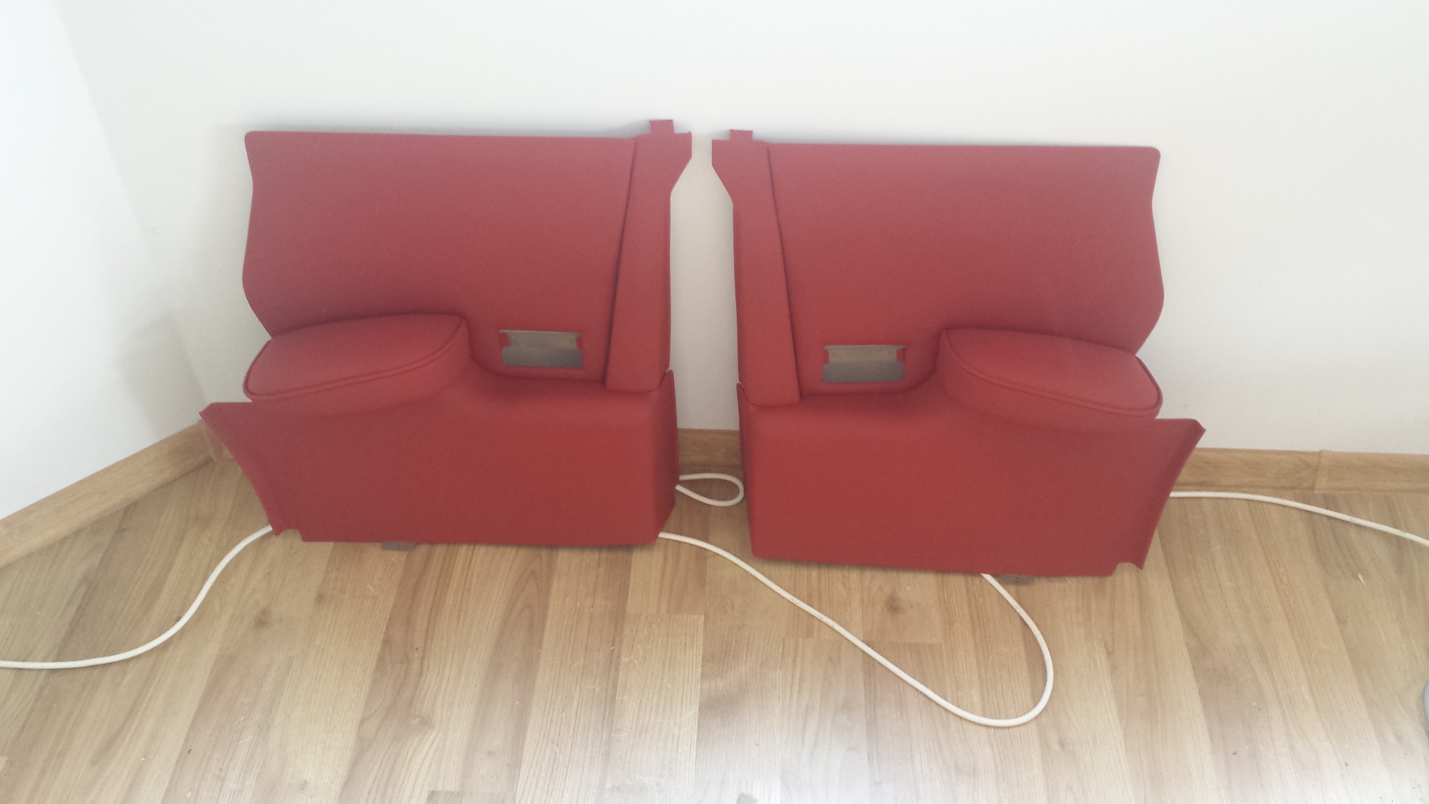 Dwa czerwone, tapicerowane panele samochodowe z widocznymi metalowymi elementami montażowymi, leżące na podłodze z parkietu, z białym kablem pomiędzy nimi.