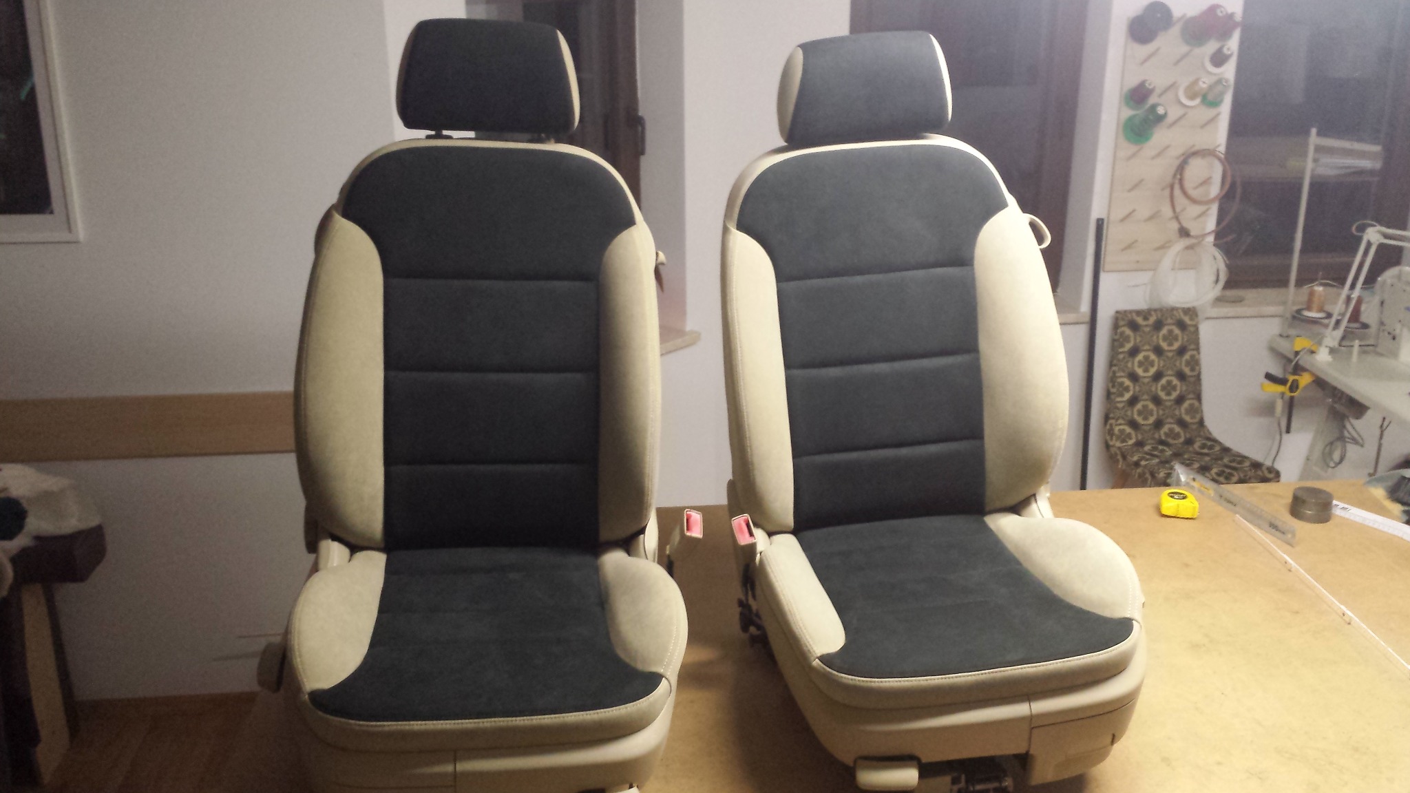 Dwa odnowione fotele samochodowe z czarną tapicerką i beżowymi bokami, widoczne różowe zapięcia pasów, na stole warsztatowym z narzędziami w tle.