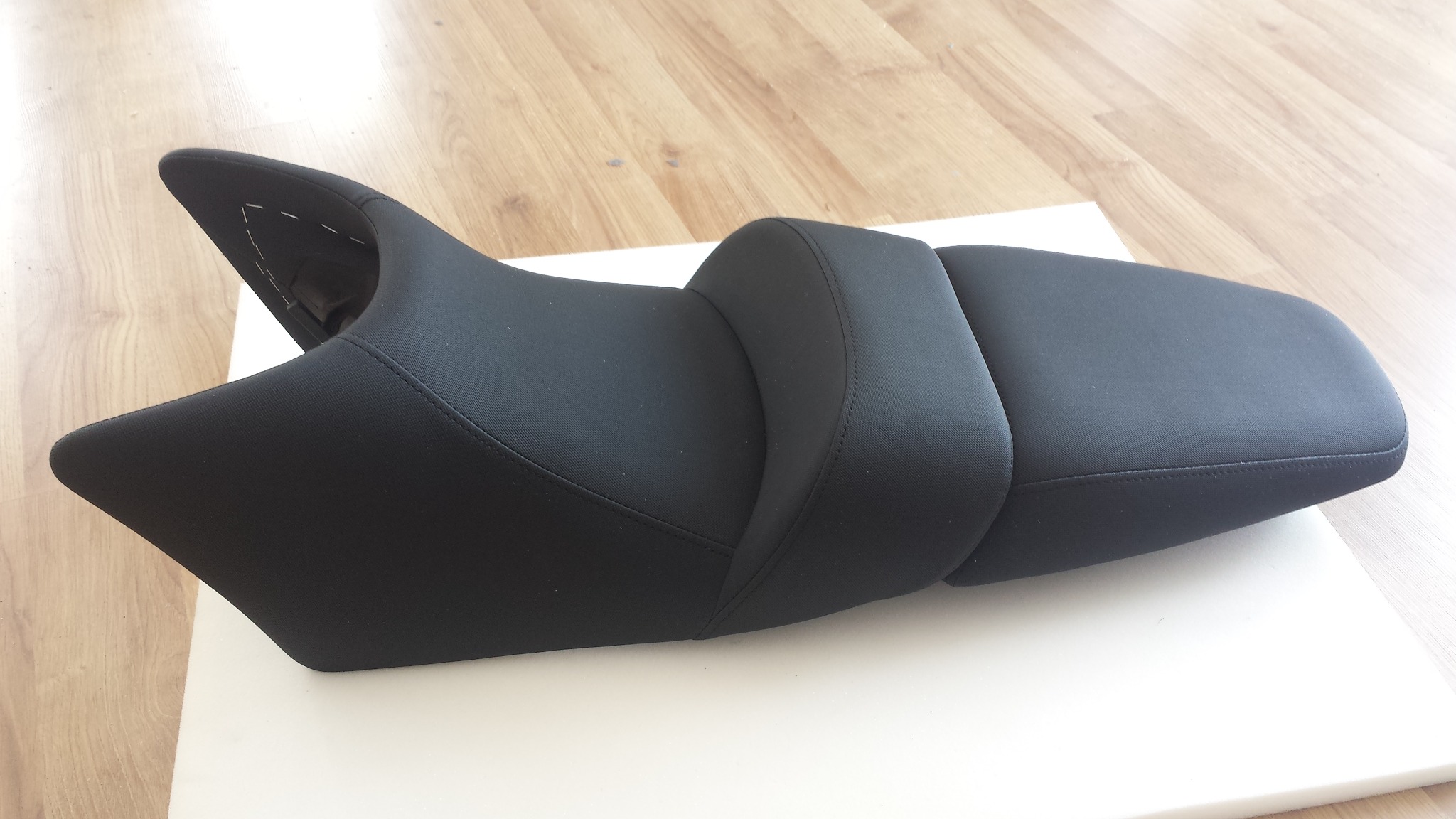 Czarne, tapicerowane siedzisko motocyklowe z wyraźnymi szwami na krawędziach, leżące na białym podłożu na tle drewnianej podłogi.