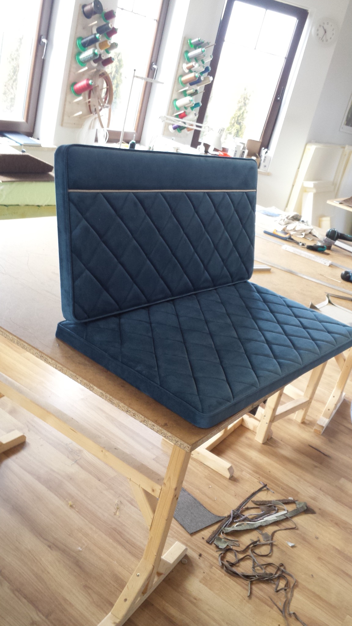 Granatowe, pikowane siedzisko z oparciem na drewnianym stole w warsztacie, widoczne szpule z nićmi przy oknie.