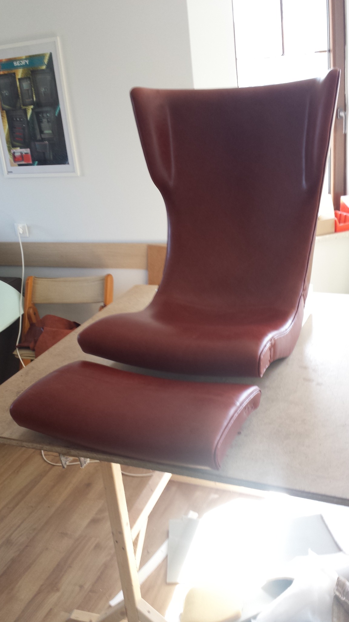 Brązowe, skórzane siedzisko i podnóżek krzesła w trakcie renowacji na warsztacie, widoczne ślady napraw i niedoskonałości.