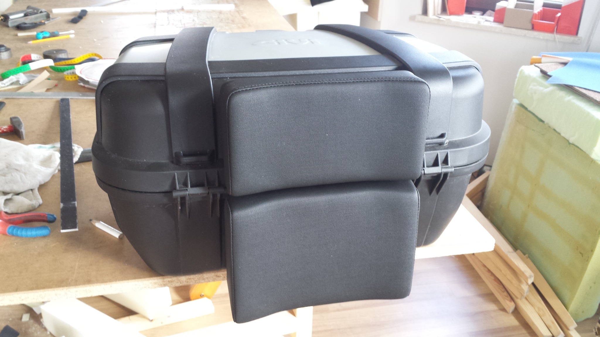 Czarny kufer motocyklowy GIVI z doczepionymi, tapicerowanymi poduszkami o prostokątnym kształcie, widoczne narzędzia i materiały tapicerskie w warsztacie.