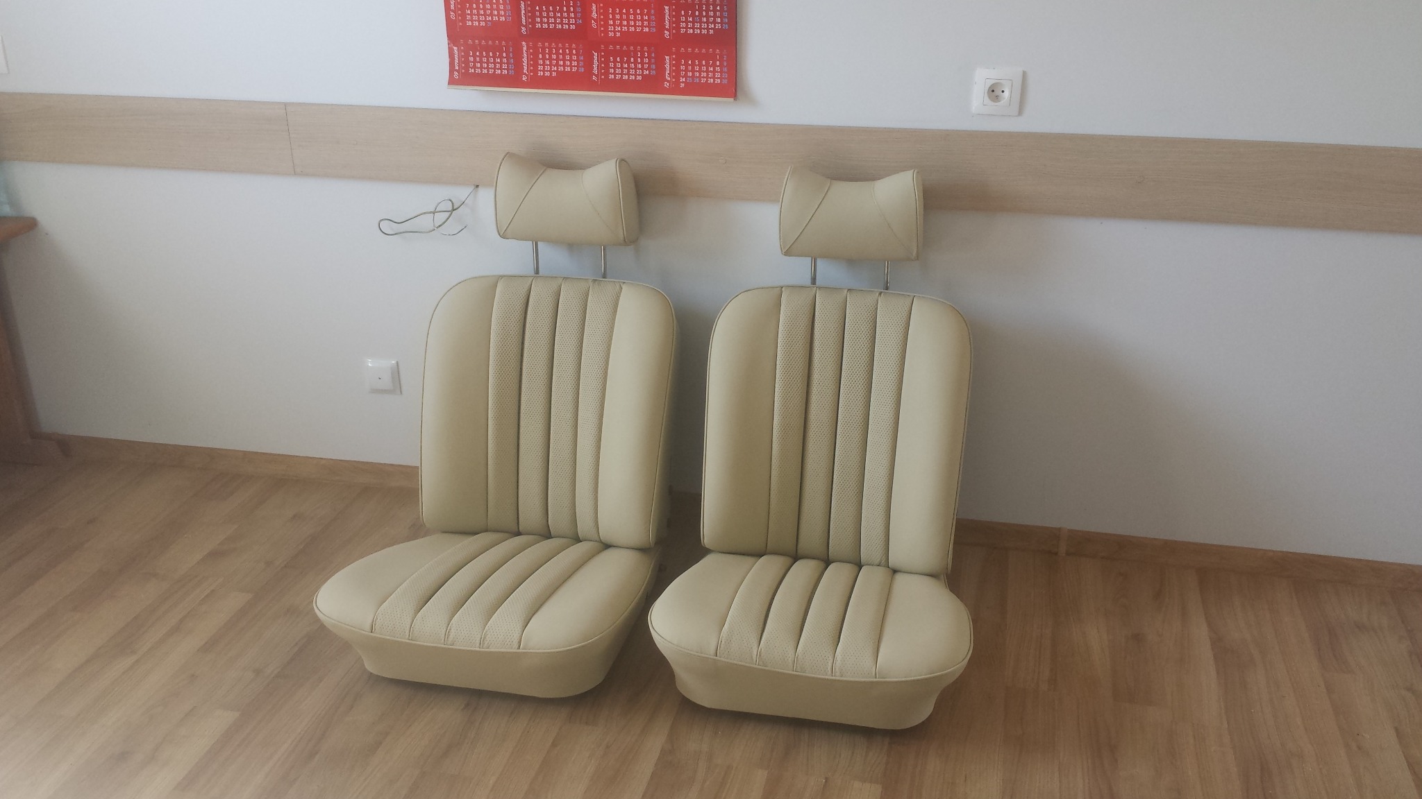 Dwa beżowe fotele samochodowe z zagłówkami, po renowacji, z perforowaną tapicerką, ustawione na podłodze z paneli.