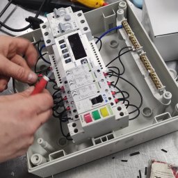 SYSTEMUS SP. Z O. O. - Elektryk podłącza przewody do rozdzielnicy wewnątrz obudowy, widoczne narzędzia i elementy instalacji elektrycznej.