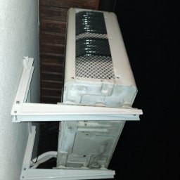 SYSTEMUS SP. Z O. O. - Zewnętrzna jednostka klimatyzatora zamontowana na białym wsporniku na ścianie budynku, widoczna od dołu, z widocznymi przewodami i kratką wentylacyjną; w tle nocne niebo i okno z ciepłym światłem.