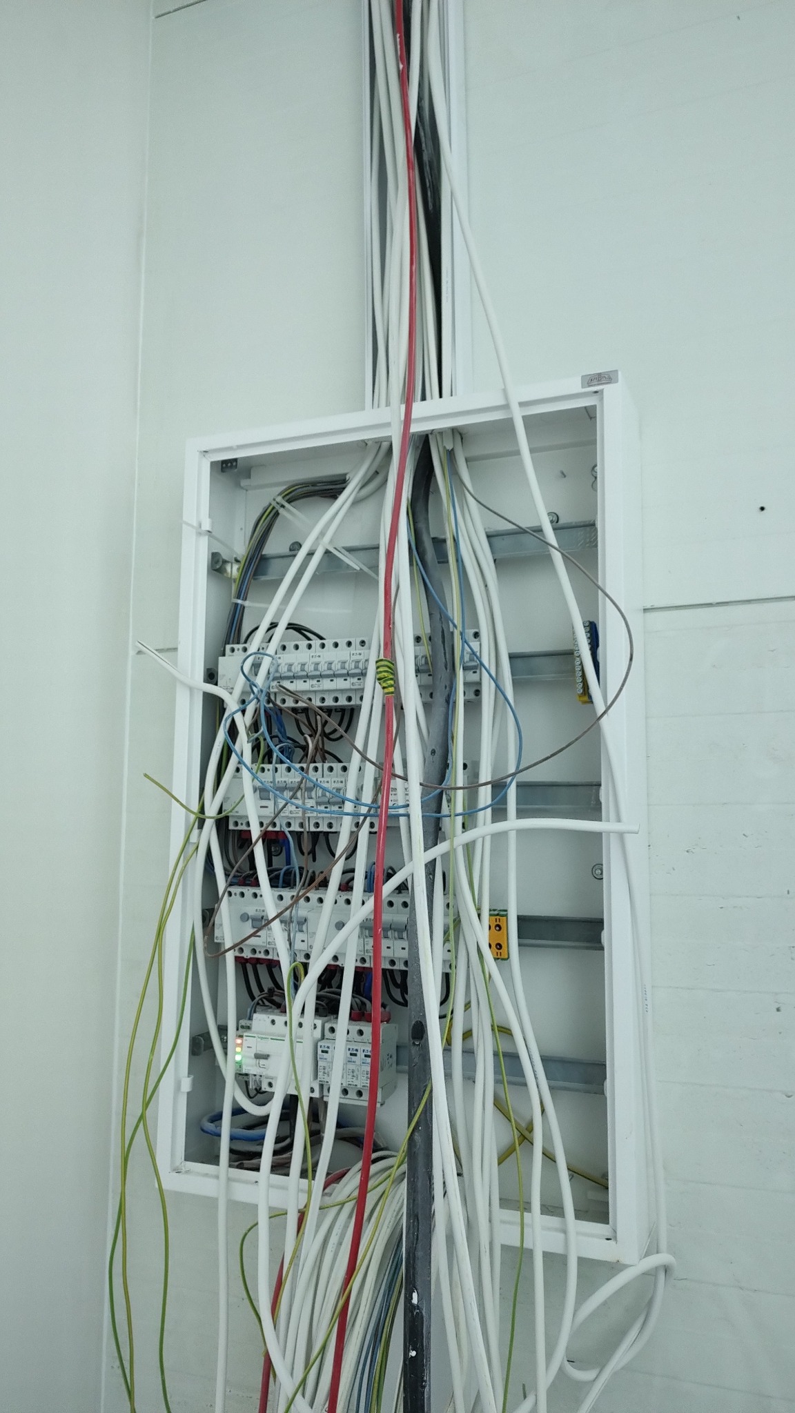 Otwarta skrzynka elektryczna z licznymi kablami wychodzącymi z góry, widoczne bezpieczniki i inne elementy instalacji, przewody w kolorach białym, czerwonym, zielonym i niebieskim.