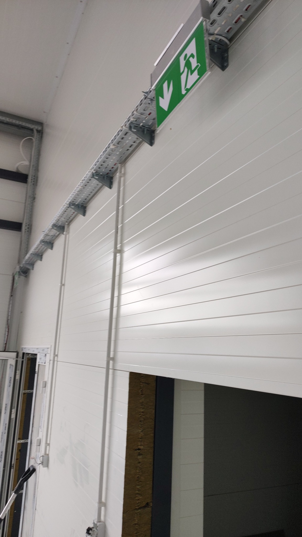 Biała ściana hali produkcyjnej z widoczną instalacją elektryczną w korytach oraz oświetleniem awaryjnym nad wyjściem ewakuacyjnym.