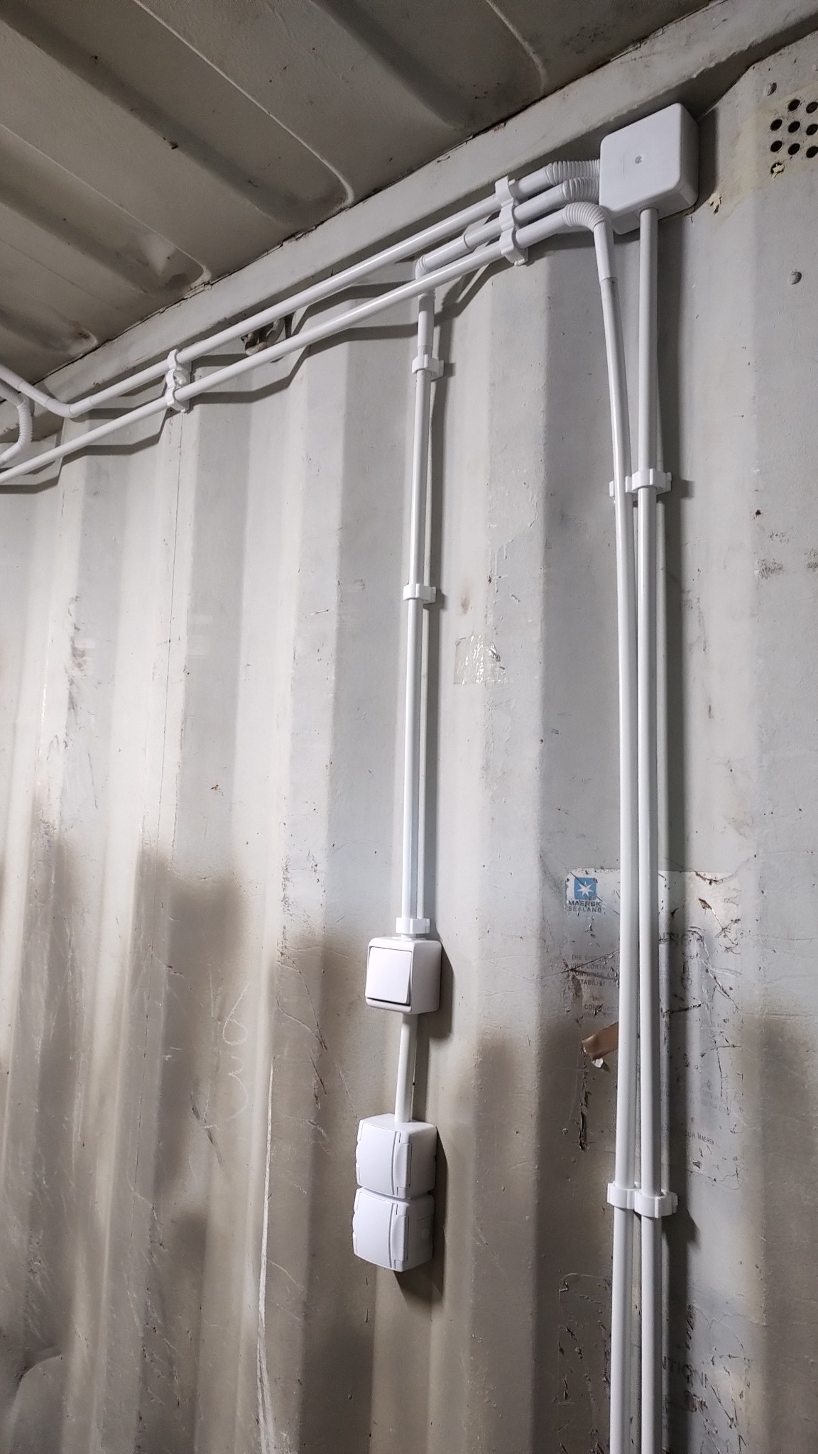 Białe rury wentylacyjne i elektryczne prowadzone po ścianie kontenera, z widocznymi puszkami instalacyjnymi i obejmami mocującymi.
