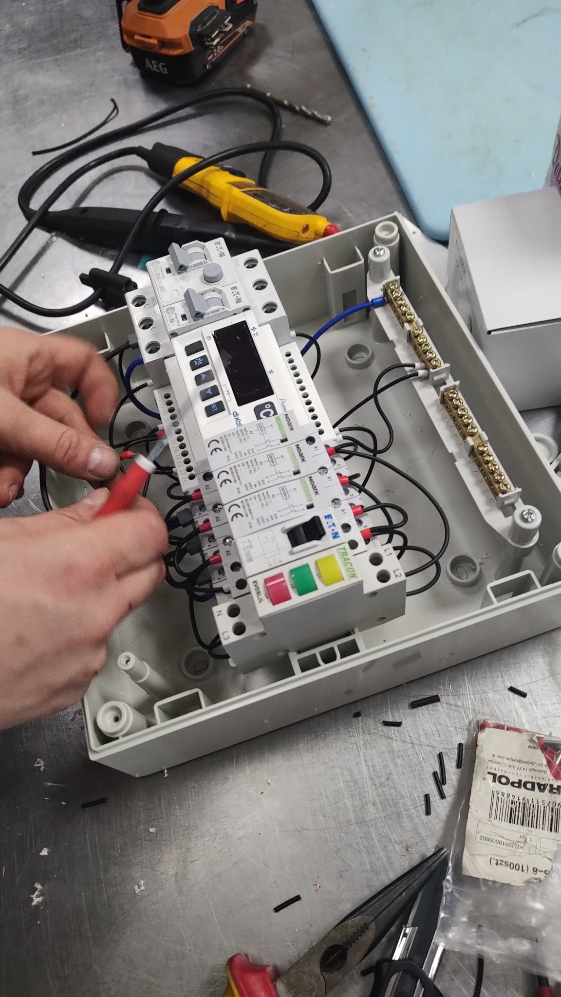 Elektryk podłącza przewody do rozdzielnicy wewnątrz obudowy, widoczne narzędzia i elementy instalacji elektrycznej.