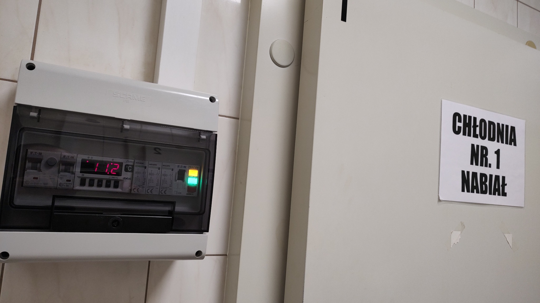 Panel sterowania chłodni z wyświetlaczem temperatury 11.2 stopni Celsjusza, zamontowany obok drzwi z napisem 'Chłodnia Nr. 1 Nabiał'.