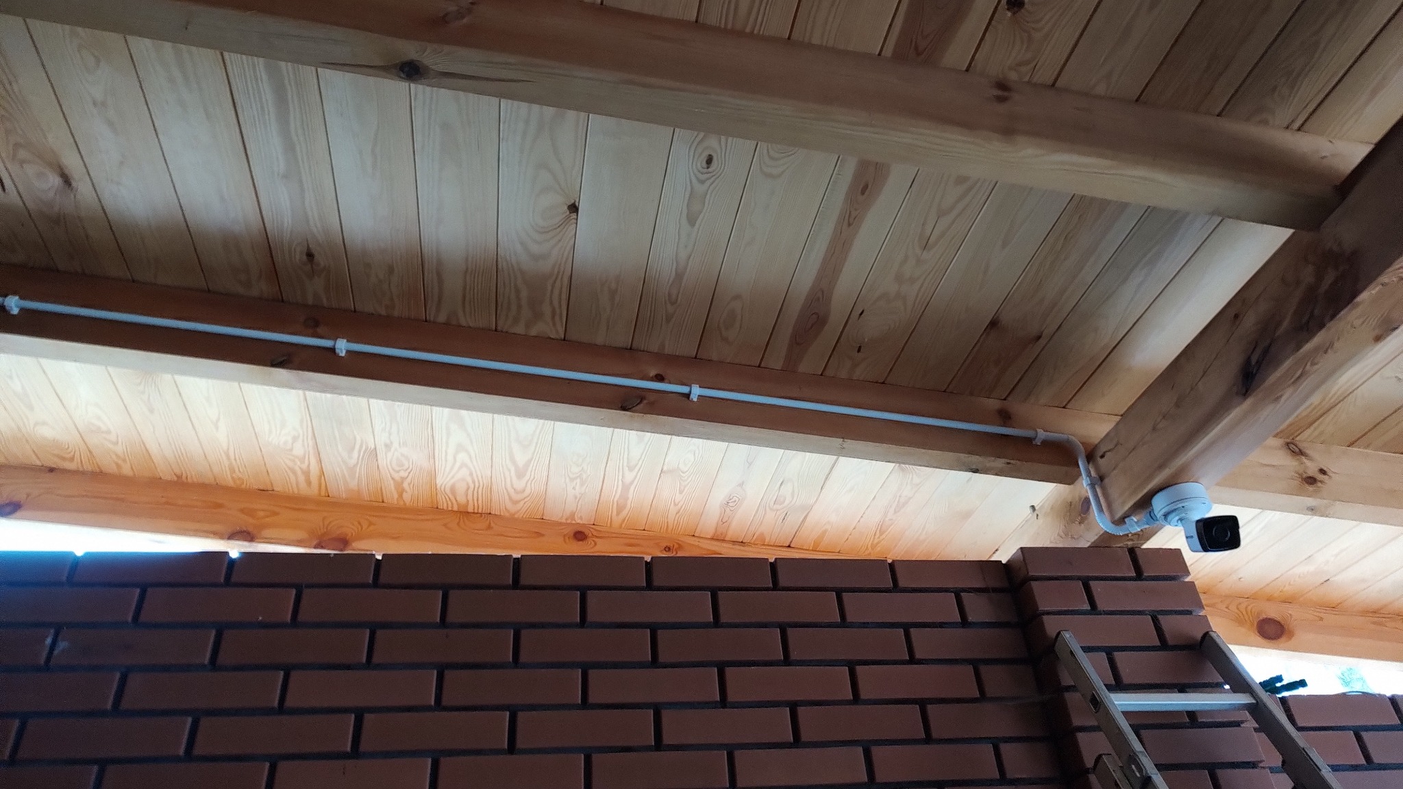 Kamera monitoringu zamontowana pod drewnianym zadaszeniem, widoczna instalacja kablowa w białej rurce, ceglana ściana w tle, drabina oparta o ścianę.