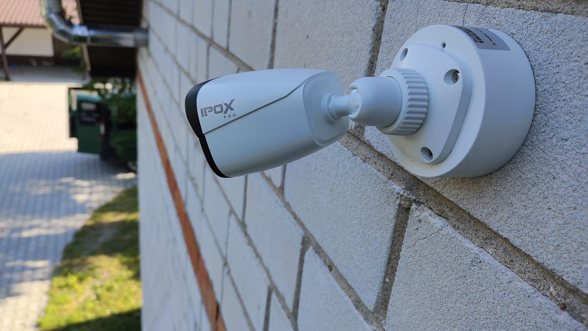Zewnętrzna kamera monitoringu IPOX Pro zamontowana na jasnej, ceglanej ścianie budynku. Widoczny fragment elewacji, rynna i zarys zieleni w tle.