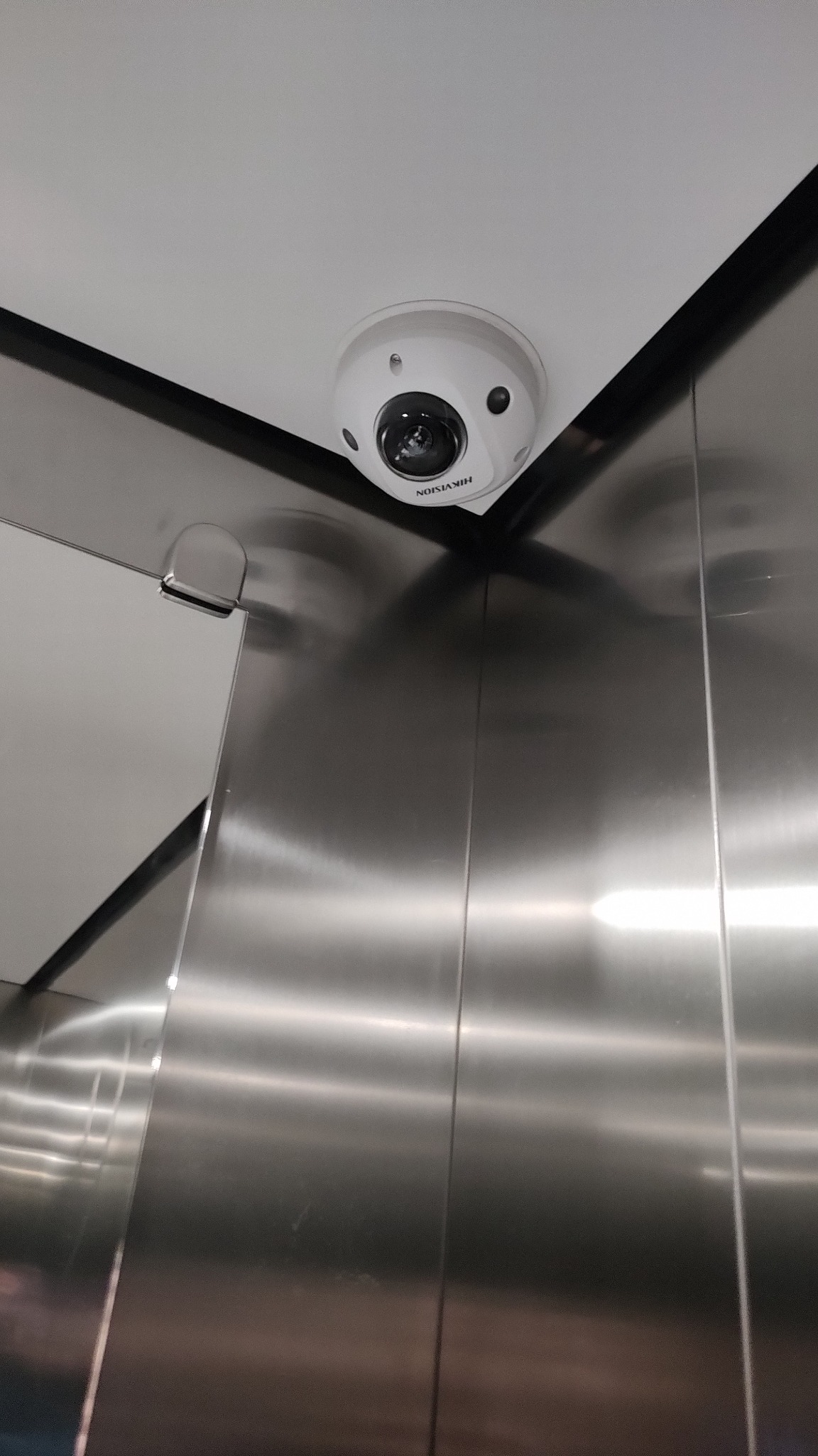 Kopułkowa kamera monitoringu Hikvision zamontowana pod sufitem w rogu windy wykończonej panelami ze stali nierdzewnej.