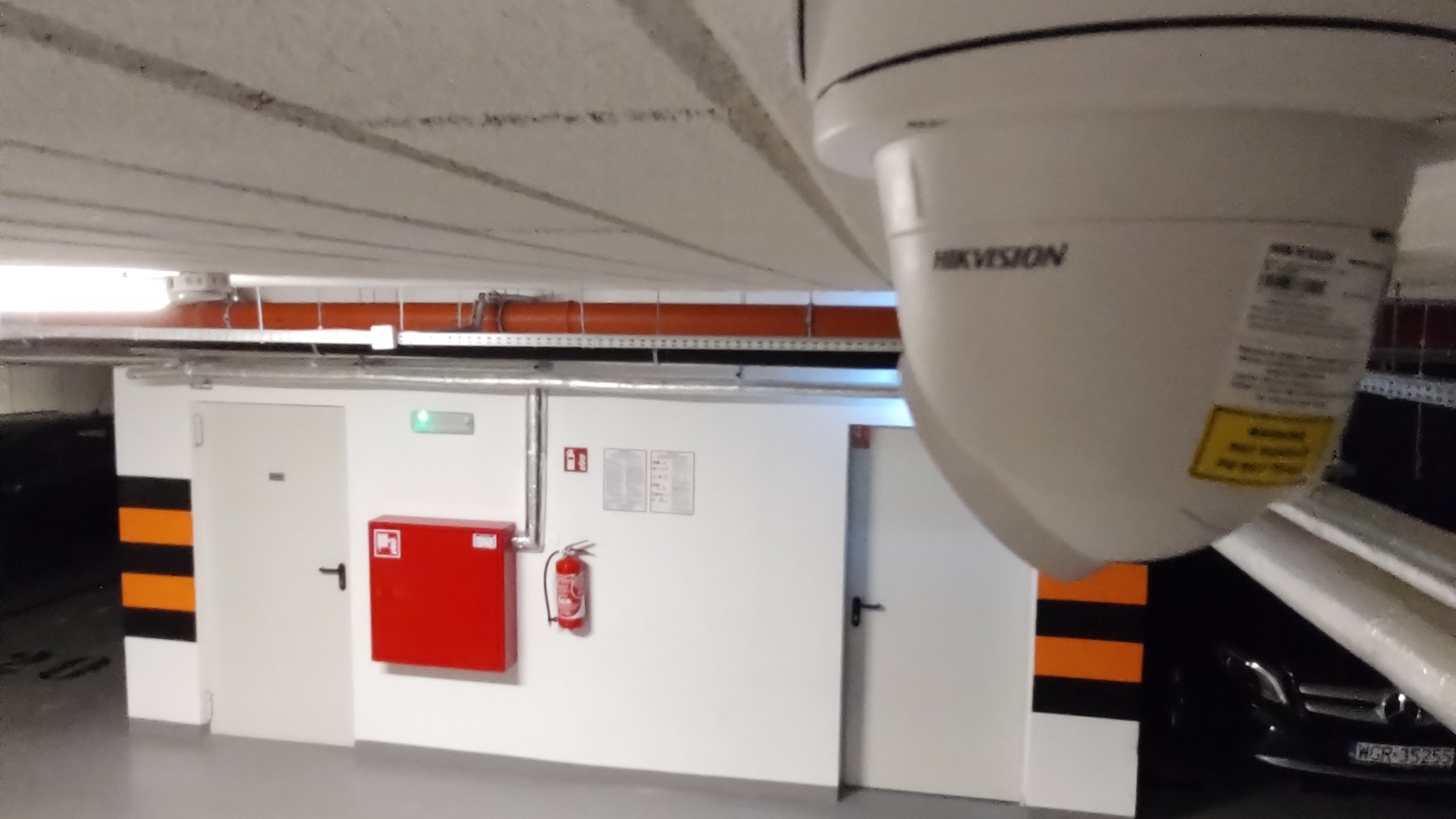 Kamera monitoringu Hikvision zamontowana pod sufitem parkingu podziemnego, widoczny fragment ściany z drzwiami, skrzynką na gaśnicę i gaśnicą.