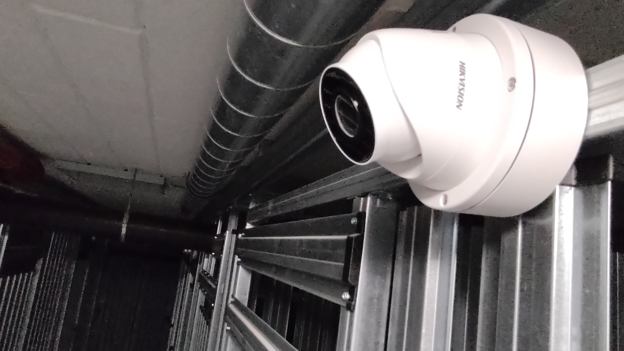 Kamera monitoringu Hikvision, zamontowana pod sufitem w pomieszczeniu technicznym, widoczna rura wentylacyjna oraz metalowa konstrukcja wsporcza.