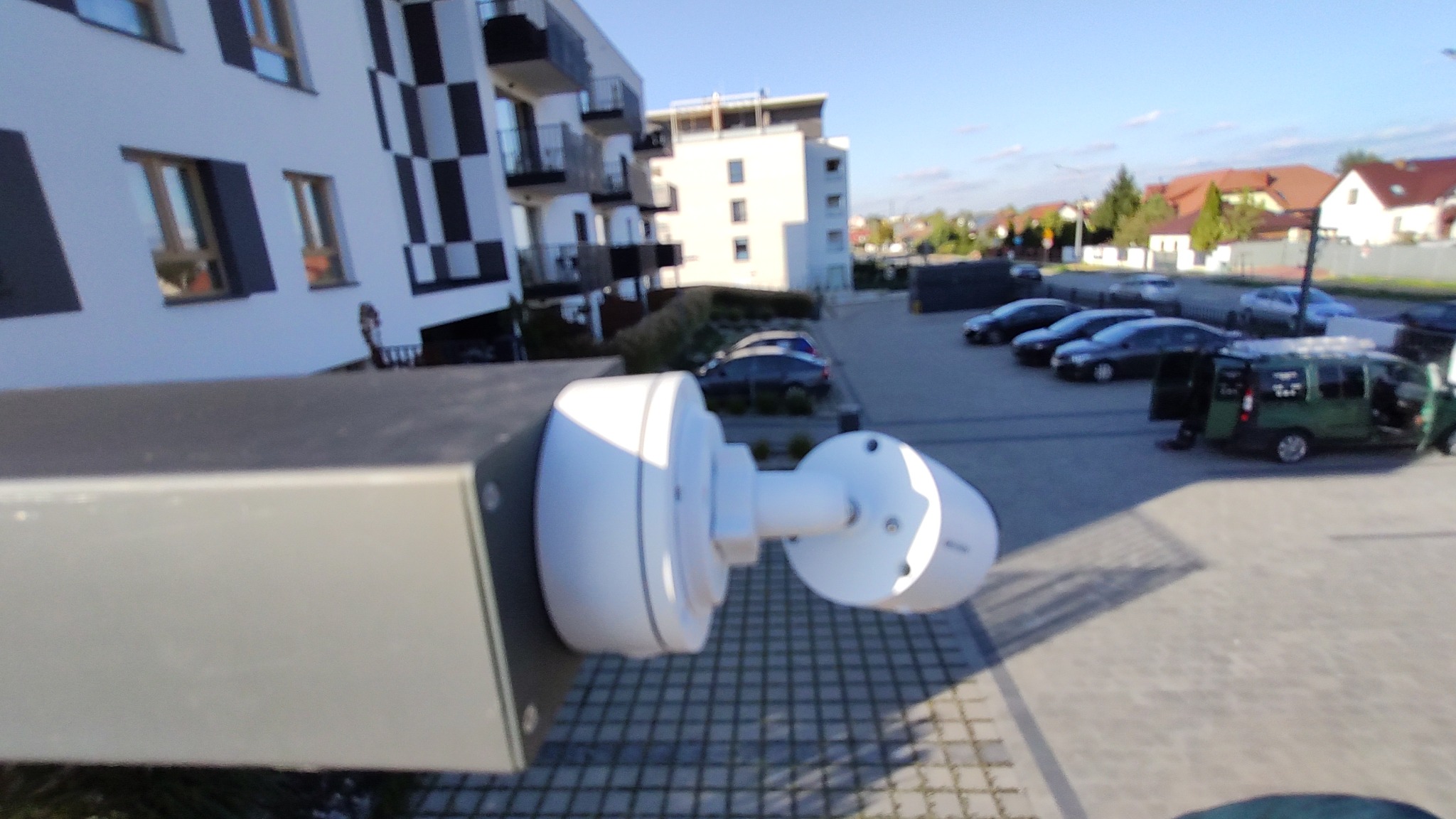 Biała kamera monitoringu zamontowana na szarym słupku, z widokiem na parking, budynki mieszkalne i błękitne niebo w tle.