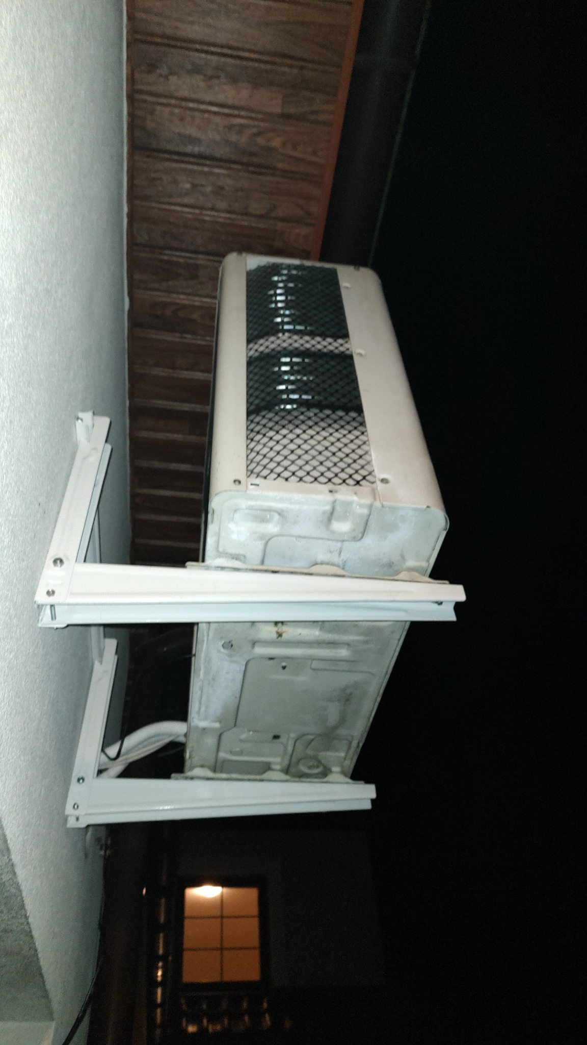 Zewnętrzna jednostka klimatyzatora zamontowana na białym wsporniku na ścianie budynku, widoczna od dołu, z widocznymi przewodami i kratką wentylacyjną; w tle nocne niebo i okno z ciepłym światłem.