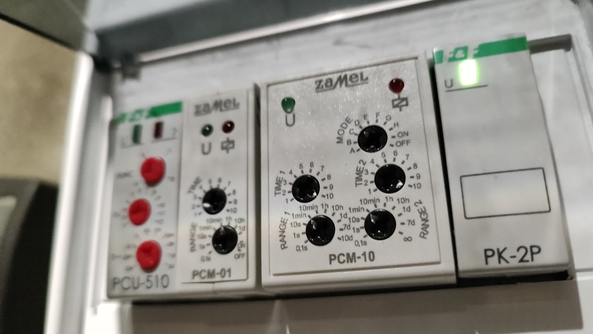 Szczegółowe zbliżenie na panel sterowania z trzema białymi modułami Zamel: PCU-510 z czerwonymi przyciskami, PCM-01 i PCM-10 z czarnymi pokrętłami do ustawiania czasu, oraz PK-2P, wszystkie...