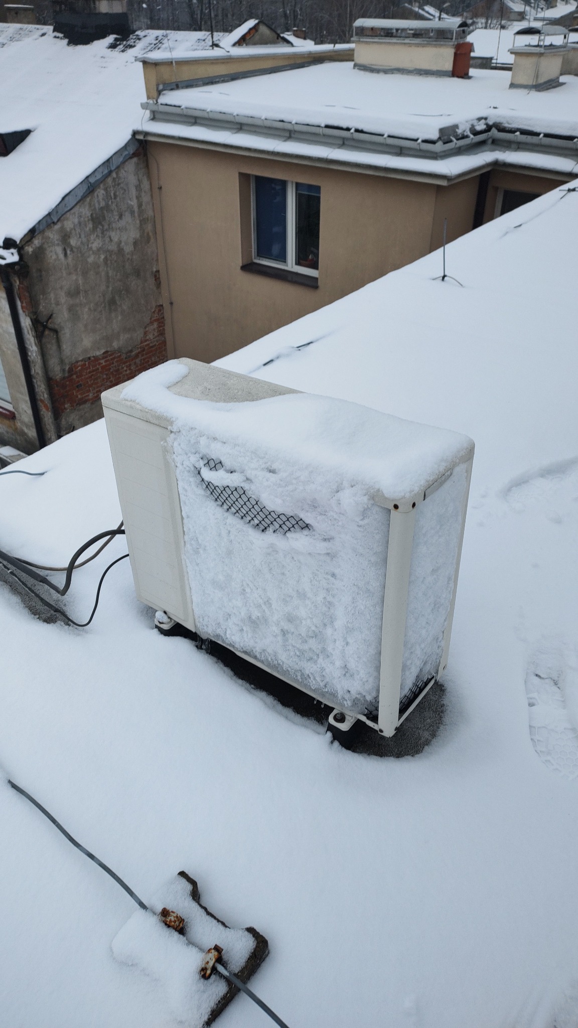 Zewnętrzna jednostka klimatyzacji na dachu budynku, częściowo zasypana śniegiem, z widoczną kratką wlotową i kablami zasilającymi.