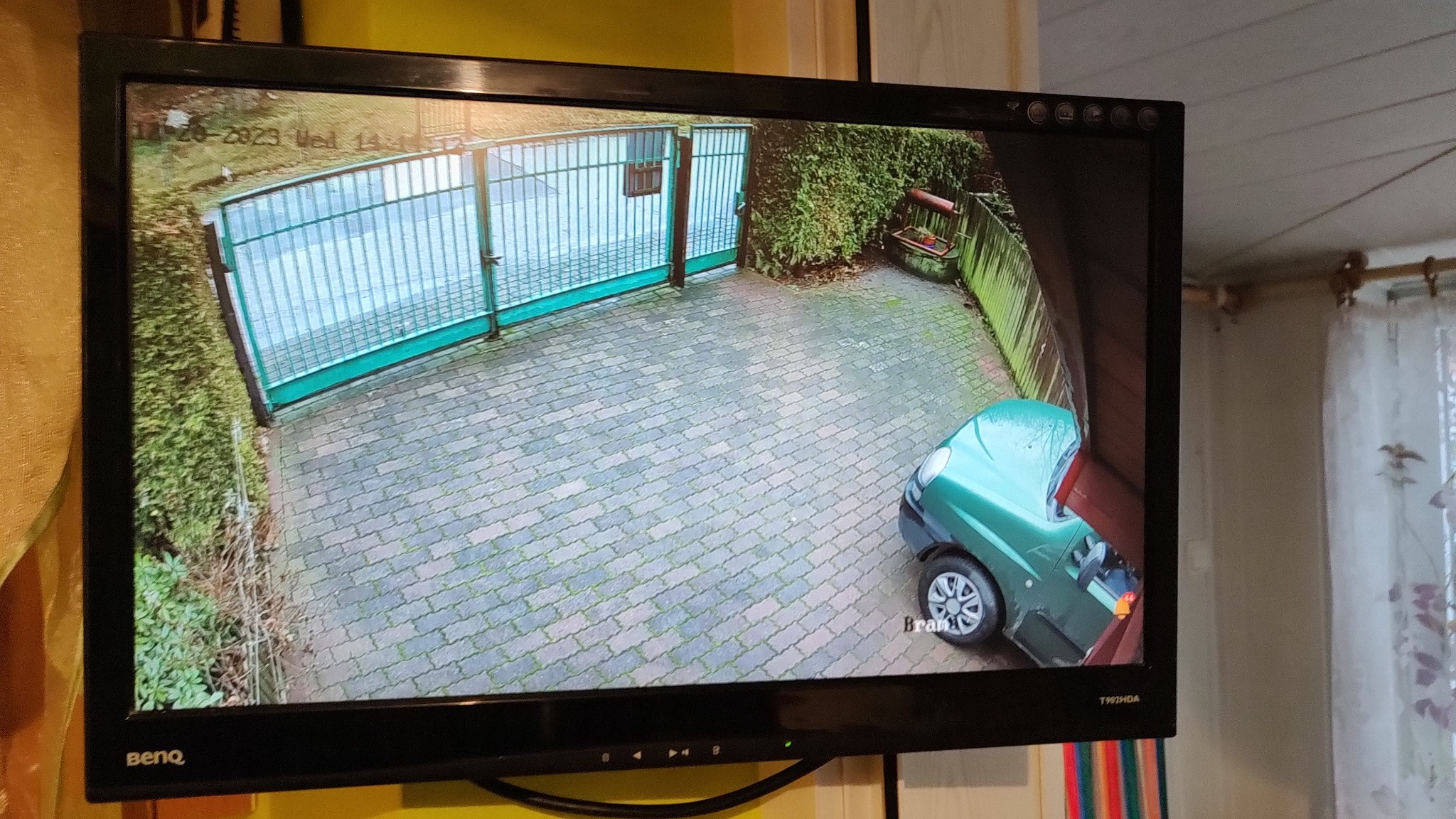 Ekran monitora BenQ pokazuje obraz z kamery monitoringu: brama wjazdowa, podjazd z kostki, zieleń i zaparkowany zielony samochód.