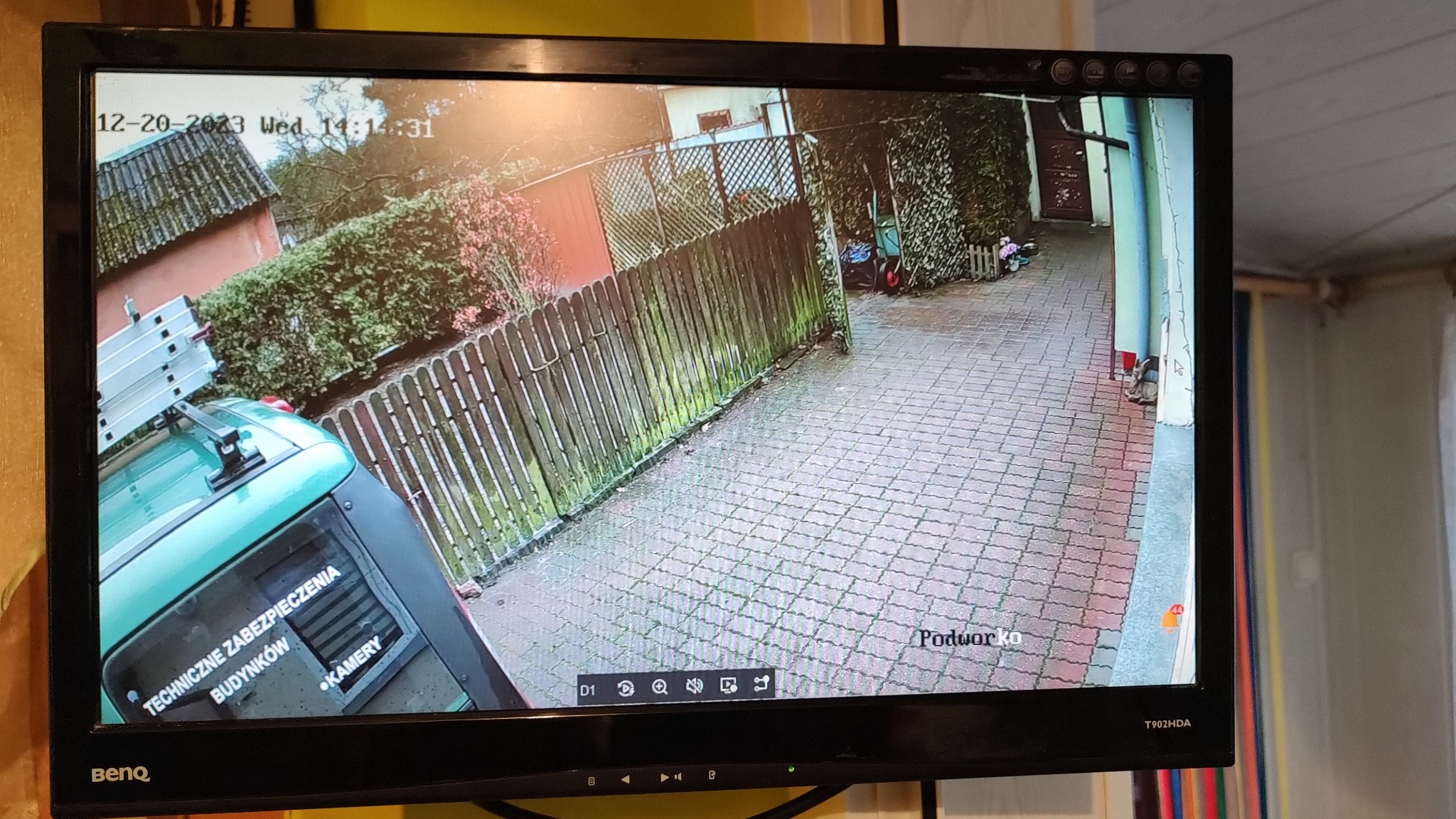 Ekran monitora BenQ z widokiem z kamery monitoringu na podwórko z zielonym samochodem firmy 'Techniczne Zabezpieczenia Budynków - Kamery', widoczny fragment dachu domu i drewniany płot.
