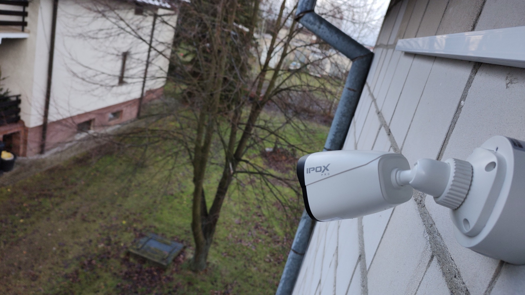 Zewnętrzna kamera monitoringu IPOX Pro zamontowana na elewacji budynku z widokiem na ogród i drzewo bez liści.