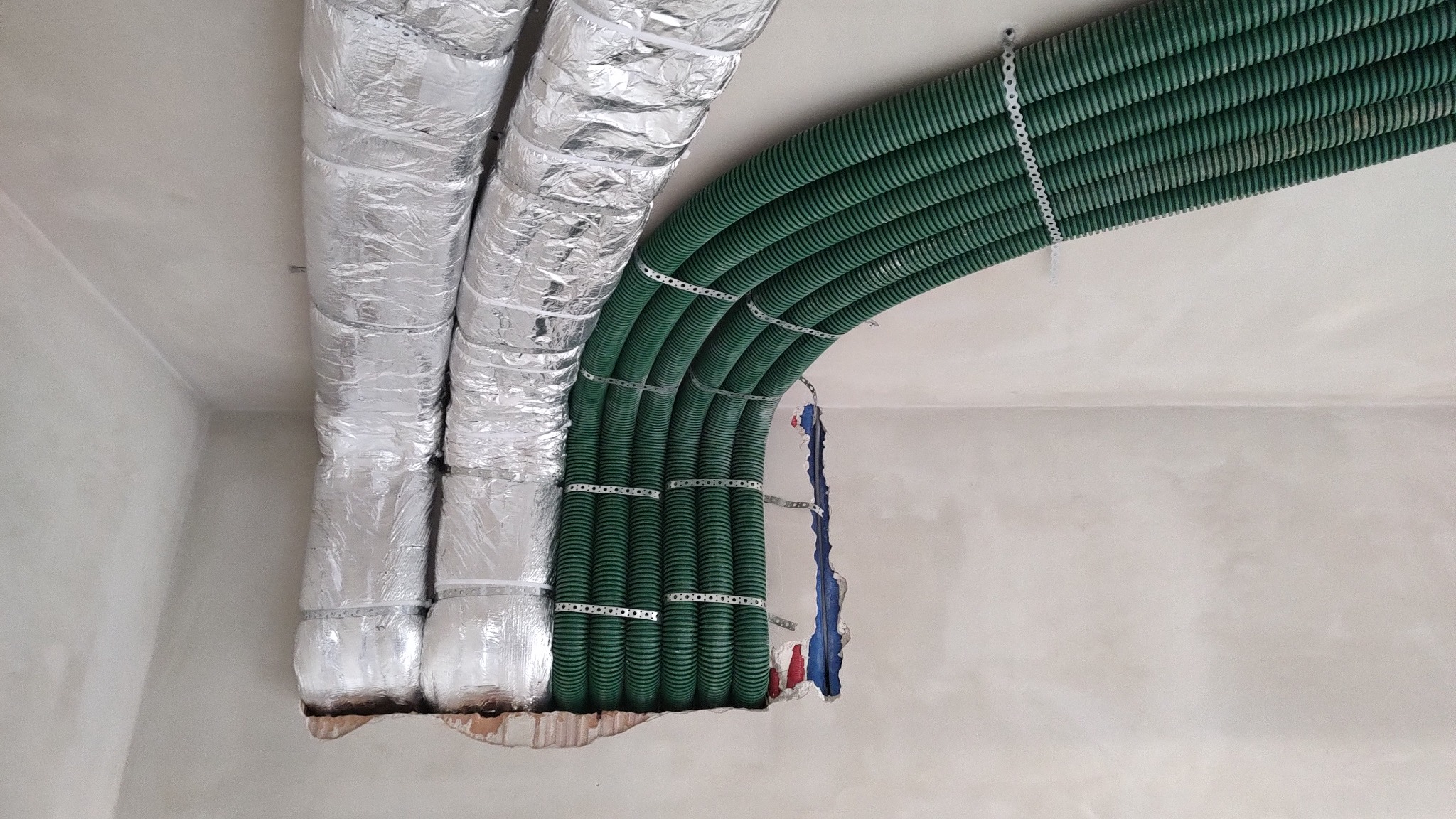 Montaż kanałów wentylacyjnych: zielone, karbowane rury przymocowane metalowymi obejmami do sufitu, obok rur w srebrnej izolacji, widoczne niedokończone prace w ścianie.