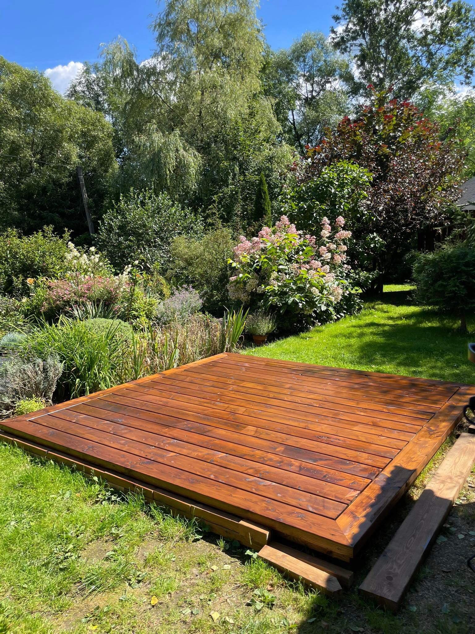 Świeżo zaimpregnowany, drewniany podest w ogrodzie w Łętowni, otoczony bujną roślinnością i trawą.