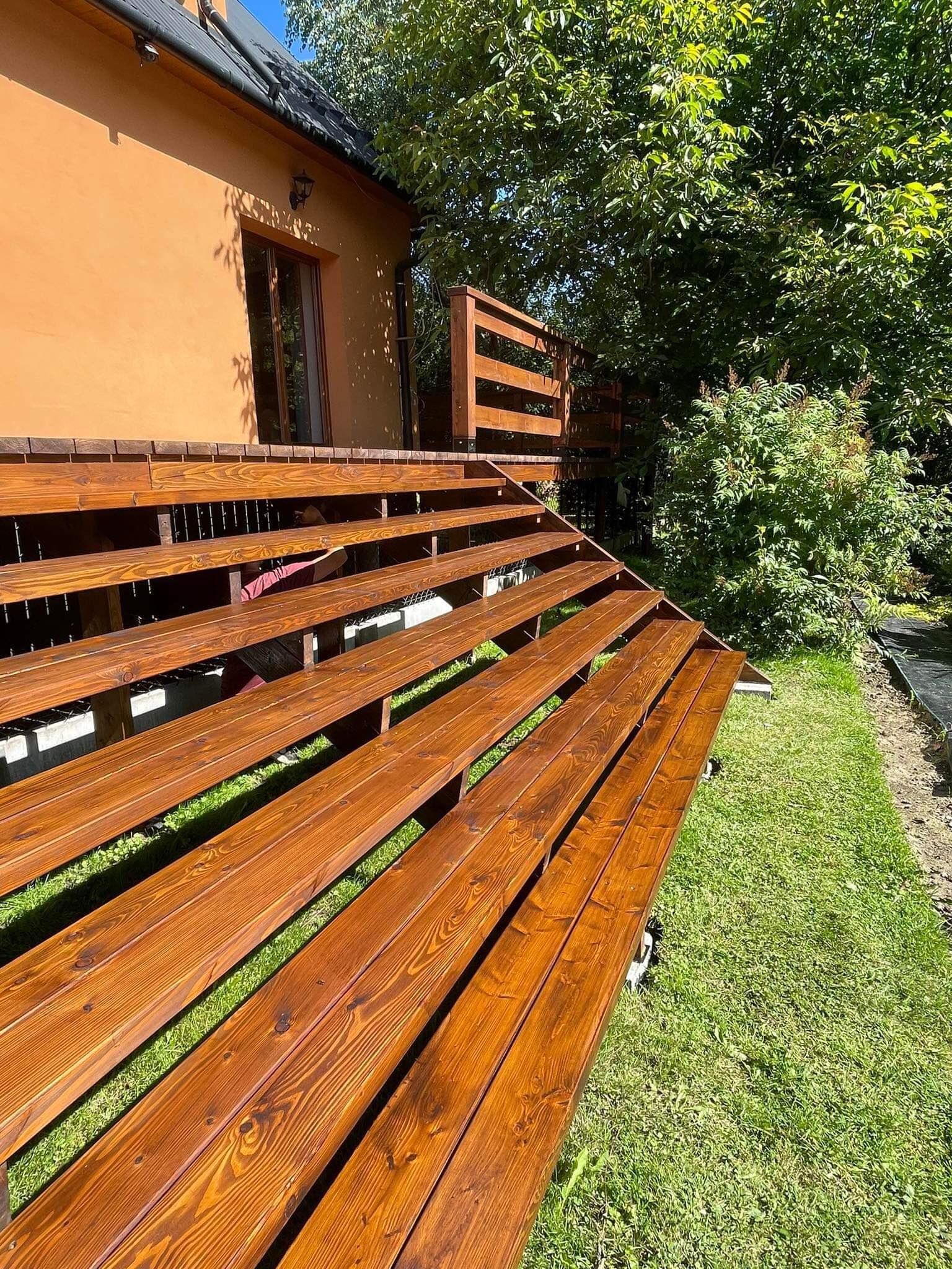 Wielopoziomowy taras z drewna o ciepłym, brązowym odcieniu, prowadzący do domu z pomarańczową elewacją, otoczony zielonym trawnikiem i bujną roślinnością.