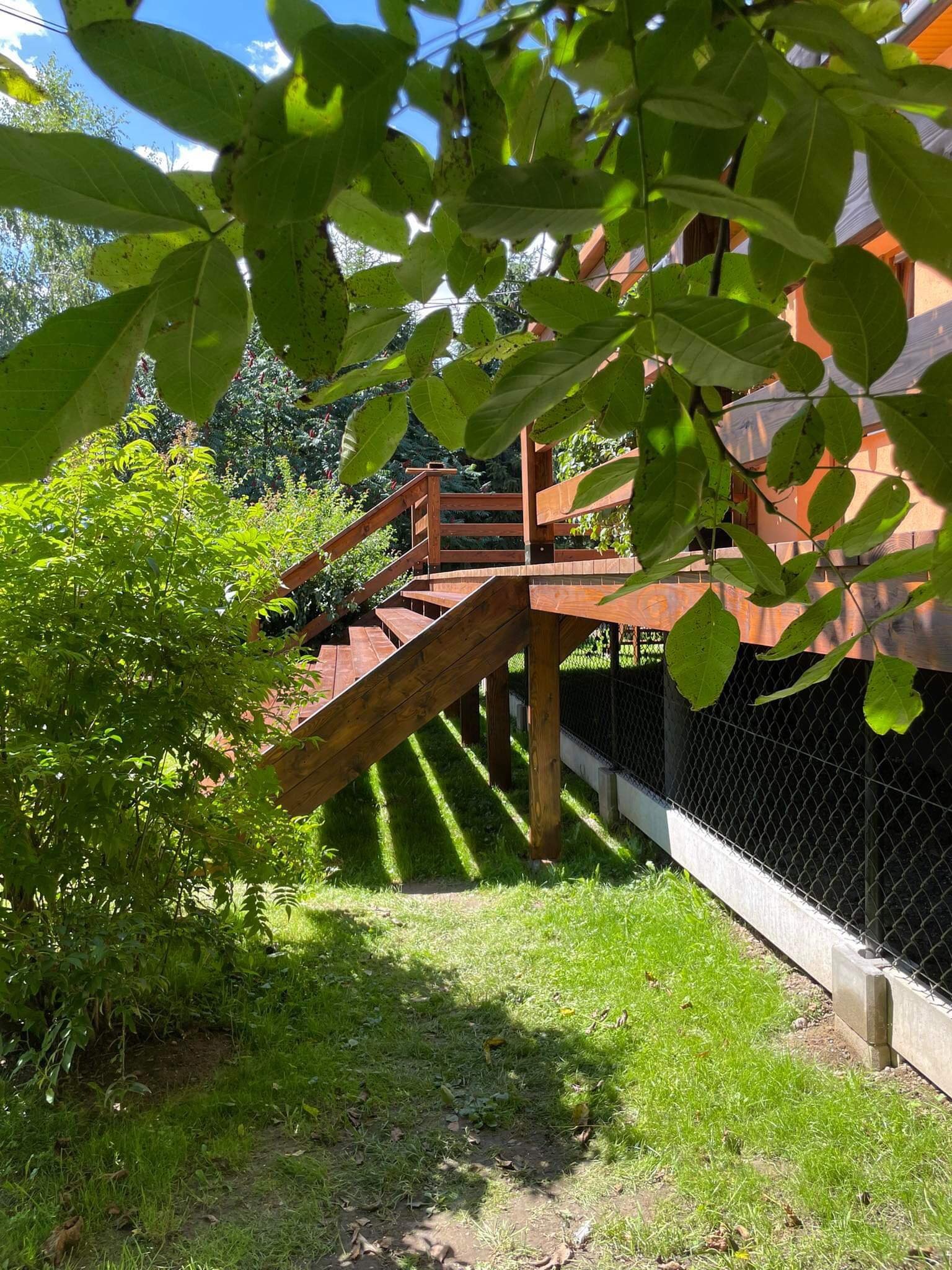 Drewniany taras z schodami, częściowo zasłonięty liśćmi drzewa, z widoczną konstrukcją pod spodem zabezpieczoną siatką, otoczony zielenią.