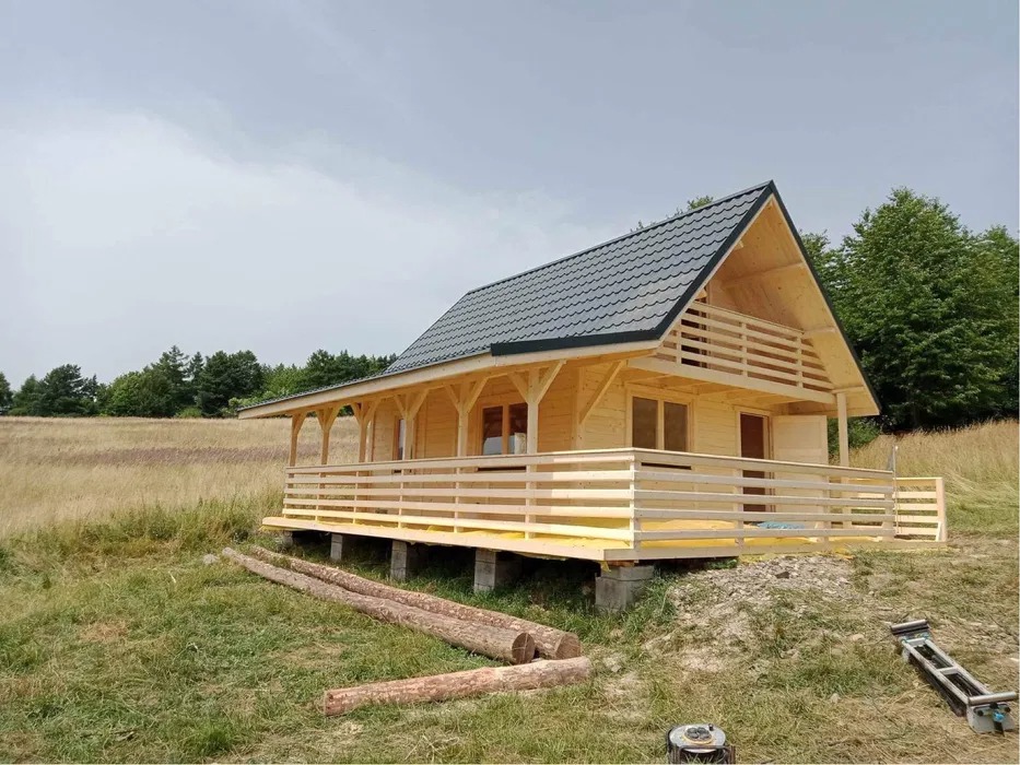 Nowo wybudowany, surowy, drewniany dom z balkonem i ciemnym dachem, położony na łące, z widocznymi elementami konstrukcyjnymi i przygotowanymi kłodami drewna obok.