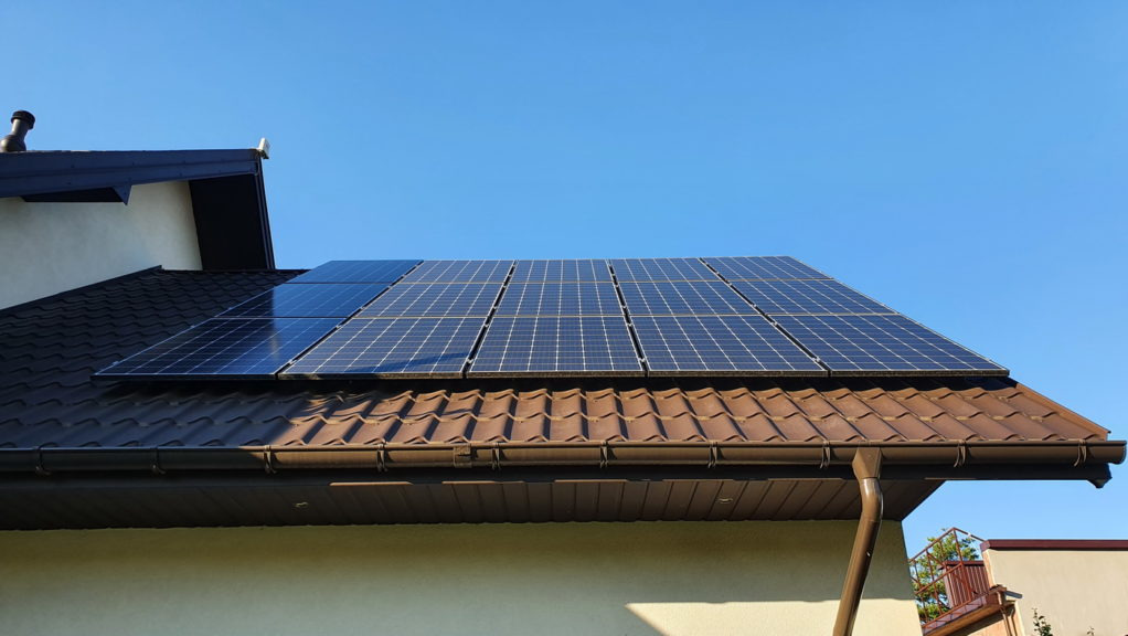 Panele słoneczne zamontowane na brązowym dachu z dachówki ceramicznej, widok z dołu na tle błękitnego nieba, rynna odprowadzająca wodę deszczową.