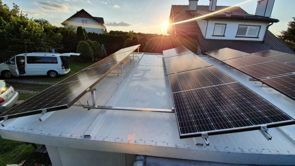 Montaż paneli słonecznych na płaskim dachu budynku mieszkalnego, widoczny biały van z otwartymi drzwiami w tle, zachodzące słońce.