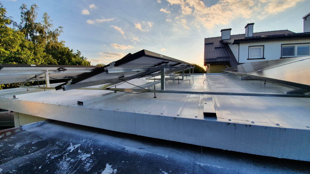 Montaż paneli fotowoltaicznych na płaskim dachu, widoczne metalowe konstrukcje wsporcze i częściowo zamontowane panele, w tle budynek mieszkalny i drzewa, zachodzące słońce.