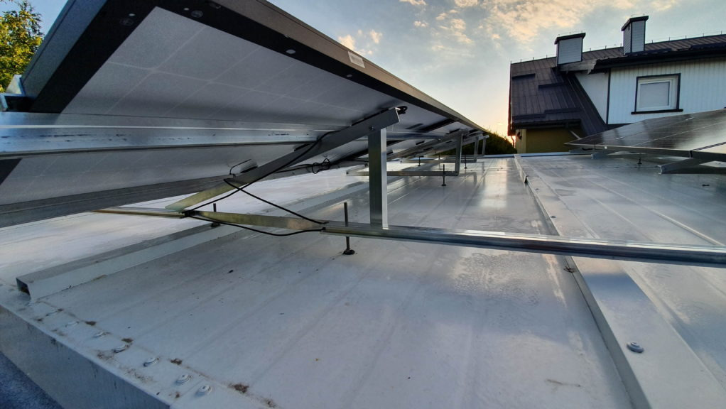 Montaż paneli słonecznych na metalowym dachu, widok od spodu, widoczne elementy konstrukcyjne i okablowanie, w tle dom jednorodzinny i zachodzące słońce.