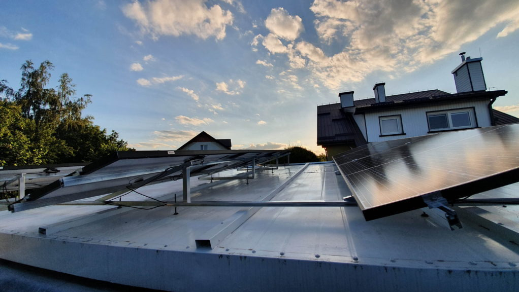 Montaż paneli słonecznych na dachu z blachy trapezowej, widoczne metalowe konstrukcje wsporcze, w tle budynek mieszkalny i błękitne niebo z chmurami oświetlone zachodzącym słońcem.