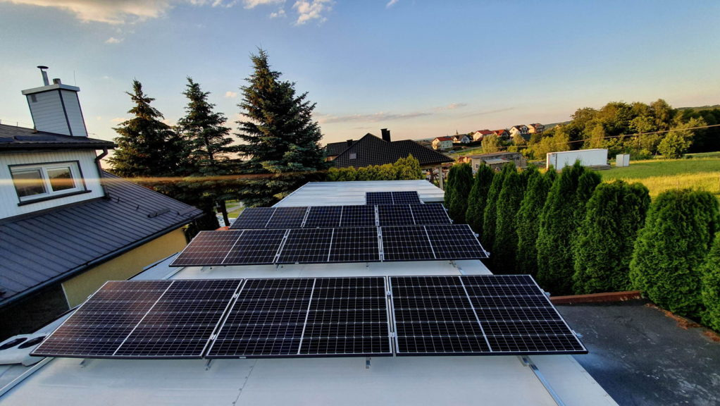 Panele fotowoltaiczne zamontowane na płaskim dachu, z widokiem na okoliczne domy i drzewa iglaste w ciepłym świetle zachodzącego słońca.
