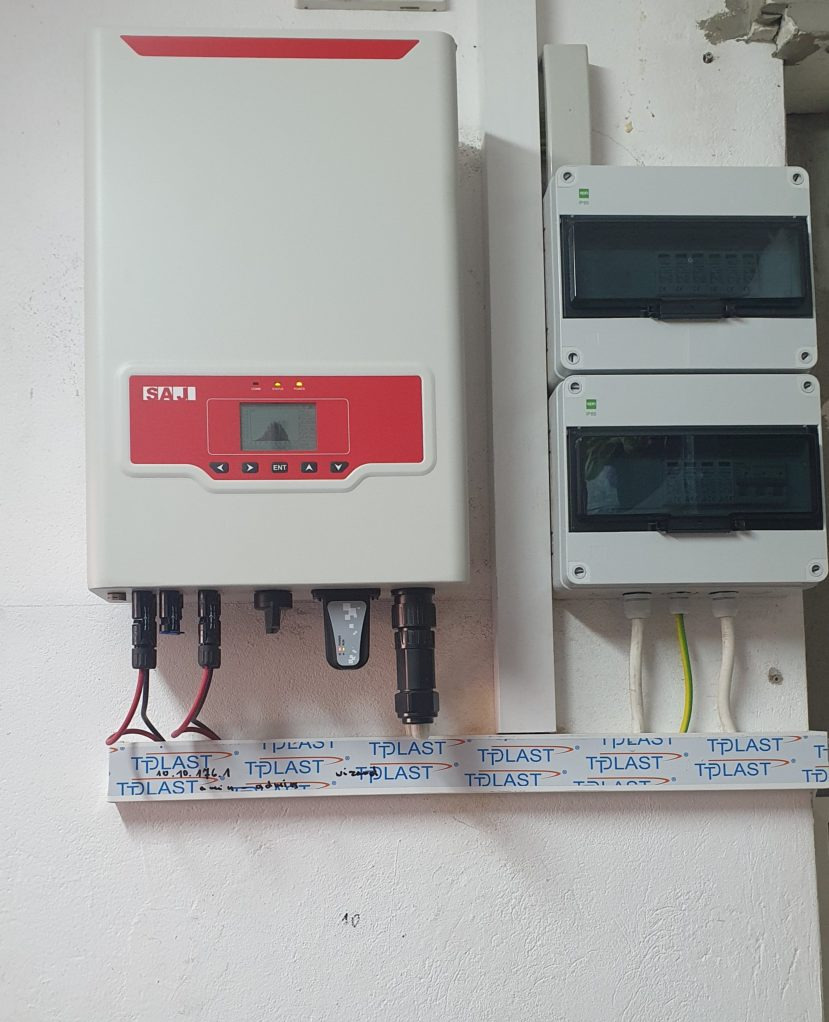 Biały inwerter fotowoltaiczny marki SAJ z czerwonym panelem sterowania, zamontowany na ścianie obok dwóch szarych skrzynek elektrycznych z przezroczystymi drzwiczkami, kable poprowadzone w listwie...