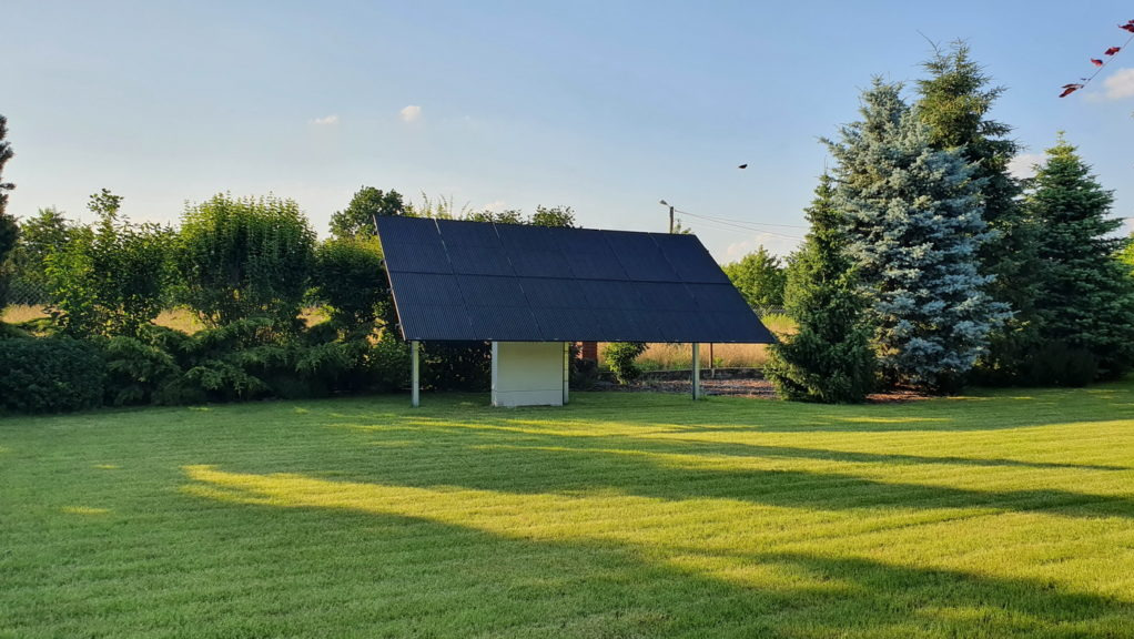 Wolnostojąca instalacja fotowoltaiczna na trawniku w ogrodzie, panele w kolorze grafitowym, pod spodem biała obudowa, w tle drzewa iglaste i krzewy.