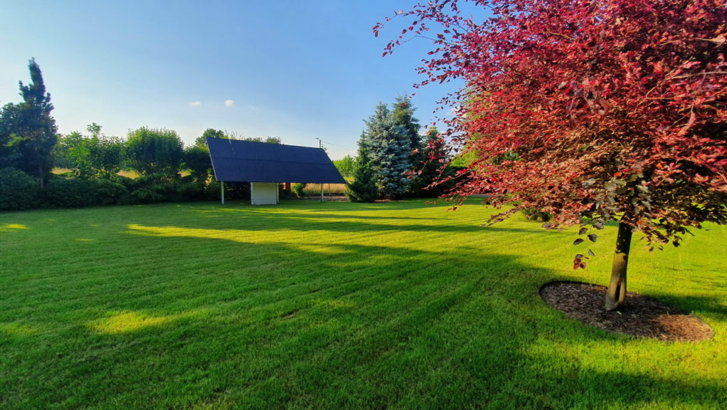 Nowoczesna instalacja fotowoltaiczna na zadaszeniu w ogrodzie, widok na zielony trawnik, drzewa i błękitne niebo w słoneczny dzień.