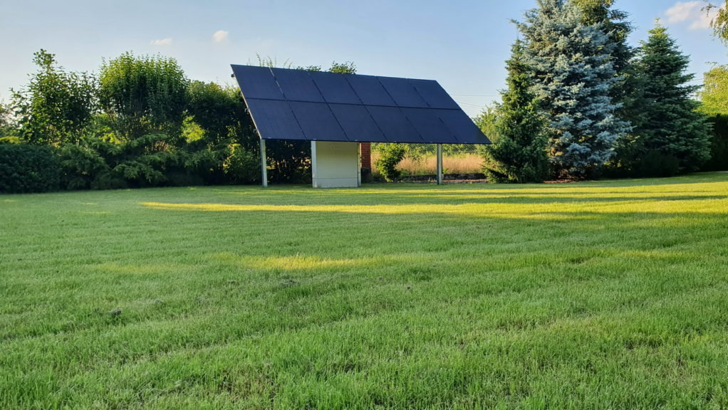 Instalacja fotowoltaiczna naziemna na metalowych słupach, umieszczona na trawniku w ogrodzie, widoczna skrzynka elektryczna pod panelami, w tle drzewa i krzewy.